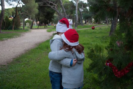 Photo for Two brothers hugging with red Santa hats next to a decorated Christmas tree. Horizontal, back view, copy space - Royalty Free Image