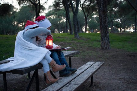 Foto de Hermano y hermana sentados en un bosque leyendo un libro de Navidad con gorra roja de santa cubierta con una manta. El invierno. Horizontal. Sin rostro. espacio de copia - Imagen libre de derechos