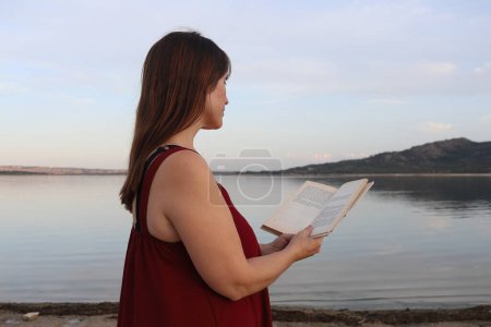 Porträt einer alleinstehenden Frau, die bei Sonnenuntergang ruhig am Ufer eines Sees ein Buch liest. Madrid. Horizontal. Kopierraum