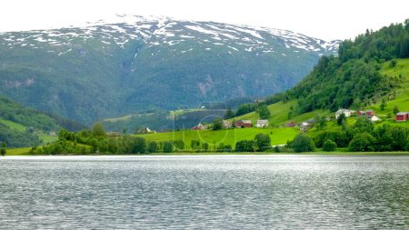 Pueblo tradicional noruego bajando la colina del fiordo rodeado de bosques verdes en las montañas cubiertas de nieve