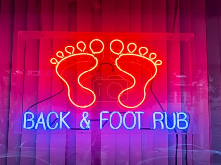 A bright red and blue neon sign illuminates the window of an anonymous massage parlor selling back rubs and foot rubs.