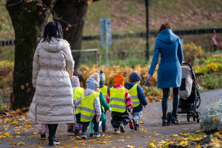 Foto de Riga, Letonia - 4 de noviembre de 2022: Un maestro de jardín de infantes con niños pequeños vestidos con chalecos reflectantes de seguridad para dar un paseo por el parque - Imagen libre de derechos