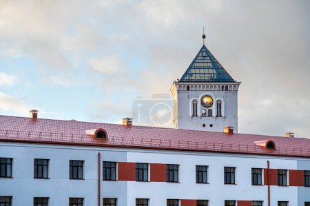 A white tower with a clock and a fragment of a modern building on the background of the sunset sky.