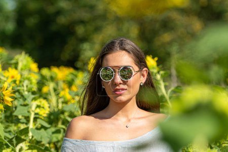 Téléchargez les photos : Femme mince sensuelle aux cheveux longs dans des lunettes de soleil dans un champ de tournesol par une journée d'été ensoleillée - en image libre de droit