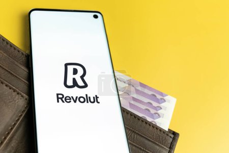 Foto de Revolut logotipo de la aplicación que se muestra en el teléfono inteligente en la cartera marrón con dinero en efectivo, fondo amarillo. SWANSEA, Reino Unido - 1 DE NOVIEMBRE DE 2022. - Imagen libre de derechos