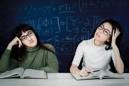 Photo for Bored female students on desk with math formula on blackboard background - Royalty Free Image