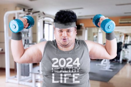 Bild eines fettleibigen Mannes hebt zwei Langhanteln im Fitnessstudio mit dem Text von 2024 gesundes Leben auf dem Hemd über verschwommenem Turnhallenhintergrund