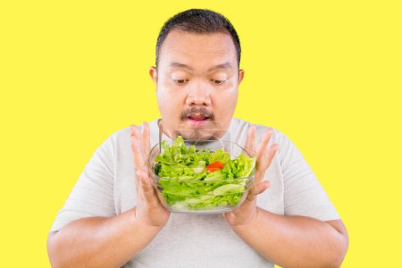 Photo for Fat Asian guy holds fresh greens salad in bowl isolated on yellow background - Royalty Free Image