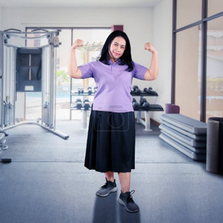 Téléchargez les photos : Surpoids femme asiatique flexion de son muscle dans une salle de musculation - en image libre de droit