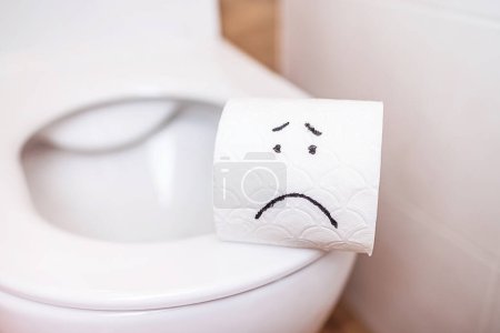 gros plan rouleau de papier toilette blanc avec un visage triste peint se tient sur les toilettes. Le concept de mauvaise humeur, problèmes de santé, constipation, hémorroïdes, problèmes intestinaux. Hygiène personnelle, autosoin
