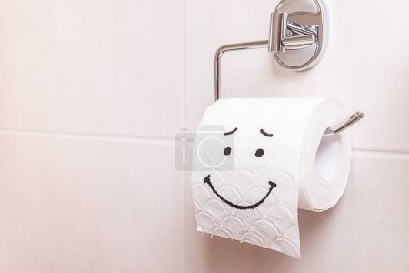 Foto de Primer plano rollo de papel higiénico blanco con una cara triste pintada se encuentra en el inodoro. El concepto de mal humor, problemas de salud, estreñimiento, hemorroides, problemas intestinales. Higiene personal, autocuidado - Imagen libre de derechos