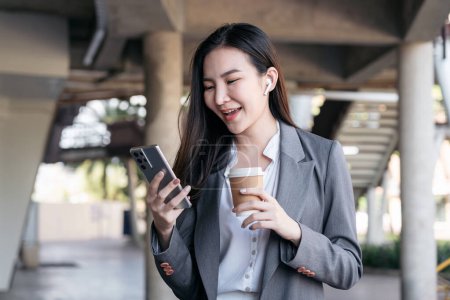 Photo for Asian businesswoman in suit, holding coffee cup to drinking and using smartphone to reading text message and typing to chatting with client while walking at outside office - Royalty Free Image