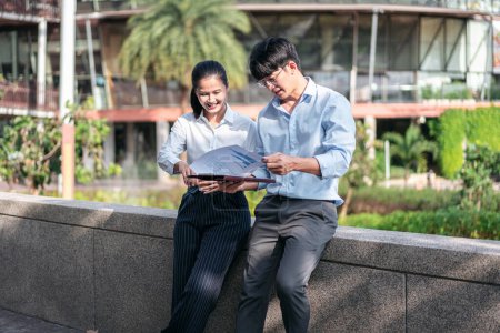 Photo for Young business man and businesswoman smiling and looking on document to checking financial report while sitting and talking about business project together at outdoor office - Royalty Free Image
