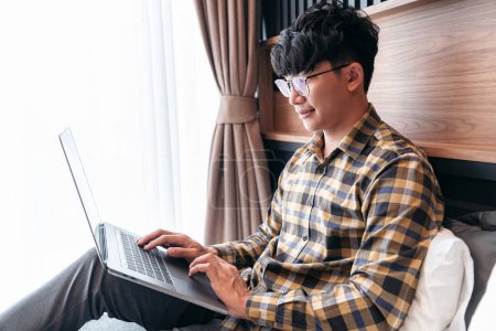 Photo for Asian man is looking on laptop screen to checking social media and typing to chatting with friend while sit lying to relaxation after working in holiday on the bed in bedroom at home - Royalty Free Image