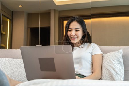 Photo for Asian woman using laptop to chatting and watching movie on social media while sitting to rest on the bed in bedroom at home and enjoy in weekend - Royalty Free Image