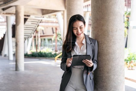 Photo for Asian businesswoman in suit, searching business data and reading text message on screen of digital tablet while standing to working at outside office - Royalty Free Image