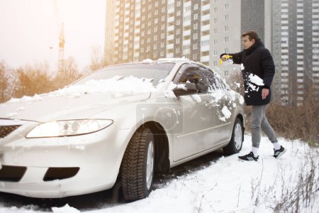 Foto de Joven limpia la nieve con un cepillo del coche, un hombre se encarga del coche en invierno - Imagen libre de derechos