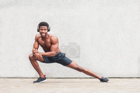 Téléchargez les photos : Un athlète afro-américain va faire du sport en plein air sans t-shirt contre un mur, un athlète dans un casque fait un échauffement, copier l'espace - en image libre de droit