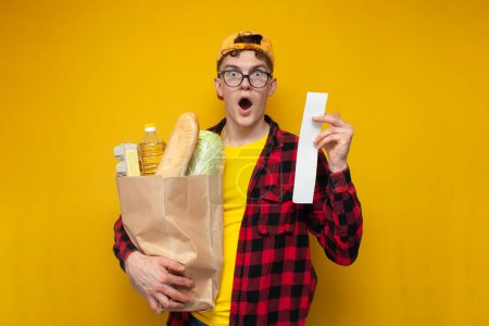 rising prices for groceries, a young guy shopper holds a package of groceries and is surprised at the high prices, a shocked shopper looks at the check on a yellow background