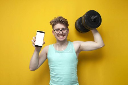 Photo for Young non-athletic guy in glasses lifts heavy dumbbells on a yellow background and shows the screen of a smartphone, a nerd trains and goes in for sports and uses the phone - Royalty Free Image