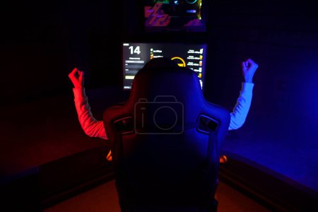 gamer in computer chair rejoice at victory at night in front of monitor, victory gesture, back view