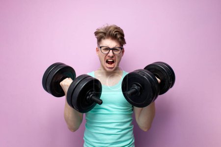 Téléchargez les photos : Young non-athletic guy in glasses holds heavy dumbbells on a pink background and trains, a serious motivated nerd goes in for sports and screaming - en image libre de droit