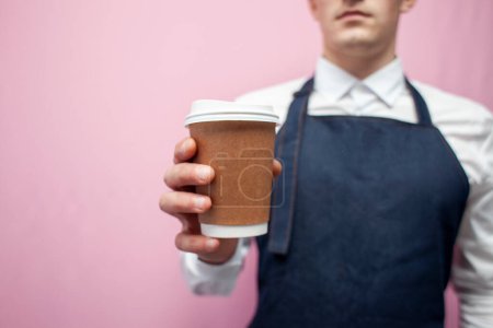 Foto de Young barista in a white shirt and apron holds a cup of coffee on a pink background, a close-up of a paper cup in the hands of a barista worker - Imagen libre de derechos