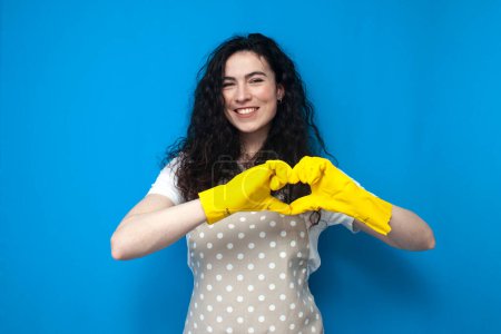 Photo for Young girl cleaner in a uniform and gloves for cleaning shows a heart with her hands and smiles on a blue background, a woman housekeeper in an apron loves work - Royalty Free Image