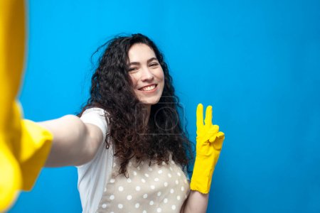Photo for Young girl cleaner in uniform and gloves for cleaning takes a selfie and shows a gesture of peace on a blue background, cleaning service, happy woman housekeeper - Royalty Free Image