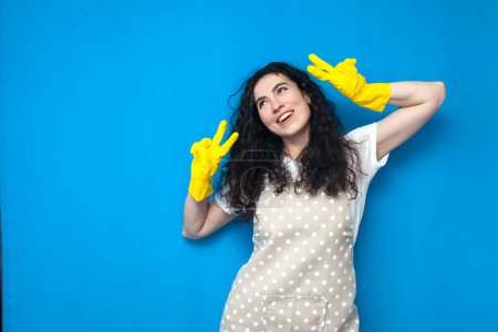 Photo for Young girl cleaner in uniform and gloves for cleaning shows a gesture of peace and poses on a blue background, happy housekeeper in an apron, cleaning service - Royalty Free Image