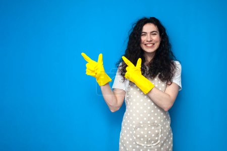Photo for Young girl cleaner in uniform and gloves for cleaning shows her hands to the side on a blue background, woman housekeeper in apron advertises copy space - Royalty Free Image