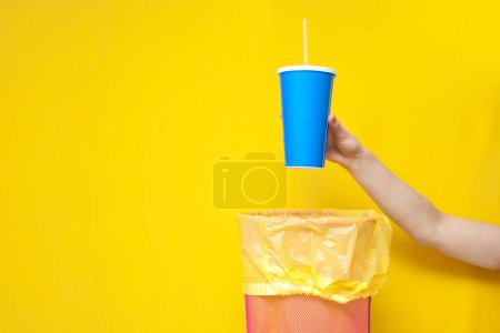 Photo for Hand throws plastic cup into trash can on yellow background, ecology concept, garbage falls into trash can - Royalty Free Image