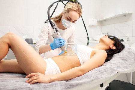 Photo for Alexandrite laser hair removal procedure in cosmetology clinic, cosmetologist doctor in uniform makes depilation of the abdomen of girl patient with modern equipment - Royalty Free Image