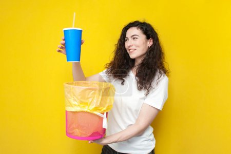Photo for Young cheerful girl throws plastic glass into a trash can, woman throws out garbage on yellow background, the concept of recycling garbage, the concept of clean ecology - Royalty Free Image