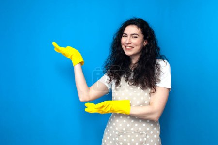 Photo for Young woman cleaner in apron and gloves for cleaning shows her hands to the side on blue background, girl housewife advertises copy space, cleaning service worker on colored background - Royalty Free Image