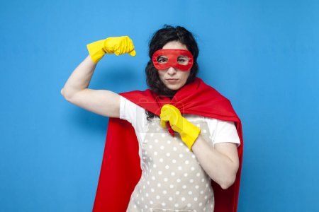Photo for Young girl cleaner in gloves and superman costume shows strength on blue background, woman housewife in superhero mask shows biceps - Royalty Free Image