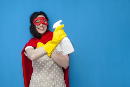 Photo for Young girl cleaner in gloves and superman costume holds detergent on blue background, woman housewife in superhero mask makes disinfection with spray - Royalty Free Image