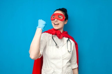 Foto de Cheerful young girl doctor in uniform in superman costume rejoice in victory on blue background, female nurse in superhero mask wins - Imagen libre de derechos