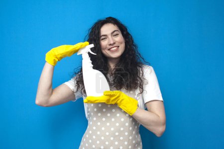 Photo for Woman housewife in gloves and apron holds detergent in her hands on blue background, woman cleaner advertises cleaning spray, cleaning service worker - Royalty Free Image