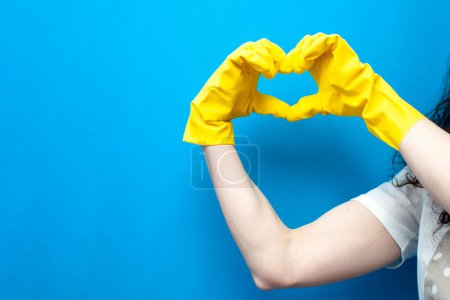 Photo for Hands in yellow cleaning gloves show heart gesture on blue background, symbol of love for cleanliness and cleaning - Royalty Free Image
