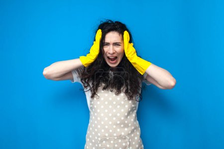 Photo for Shocked and confused woman cleaner in apron and cleaning gloves screams on blue background, housewife girl in panic and stress, cleaning service worker on colored background - Royalty Free Image