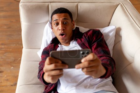 Foto de Joven sorprendido afroamericano hombre se encuentra en cómodo sofá y juega videojuegos en el teléfono inteligente, chico sorprendido jugador juega juegos móviles en casa - Imagen libre de derechos