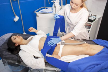 coolsculpting procedure in cosmetology clinic, girl patient lies on couch and cosmetologist doctor makes procedure to remove fat from the abdomen with modern equipment