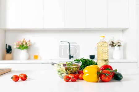 Foto de Ingredientes para ensalada y productos se encuentran sobre la mesa en la cocina moderna blanca, verduras, tomates, pimientos, botella de aceite en la cocina vacía luz, comida vegetariana saludable - Imagen libre de derechos