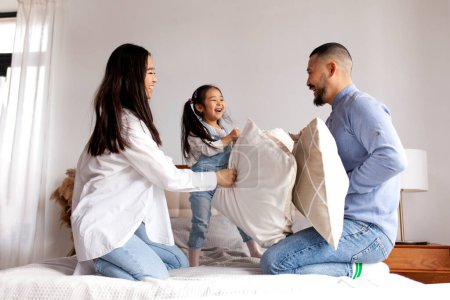 Téléchargez les photos : Bataille d'oreillers, petite fille asiatique joue avec ses parents sur le lit à la maison, les parents coréens passent du temps avec leur fille et se réjouissent, maman et papa s'amusent avec l'enfant - en image libre de droit