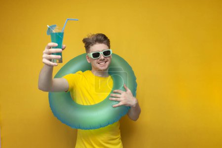 Photo for Tourist at sea. a young guy in sunglasses holds a blue refreshing cocktail and smiles on a yellow background - Royalty Free Image