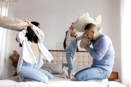 happy asian family pillow fight on the bed and rejoice, little korean girl active rest with dad and mom and hit dad with soft pillow at home, child play with parents and rejoice