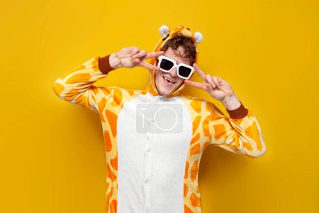 Foto de Joven alegre chico en divertido bebé jirafa pijamas y gafas está bailando sobre fondo amarillo, hombre en animal cosplay ropa muestra gesto de paz, concepto de fiesta de pijama - Imagen libre de derechos