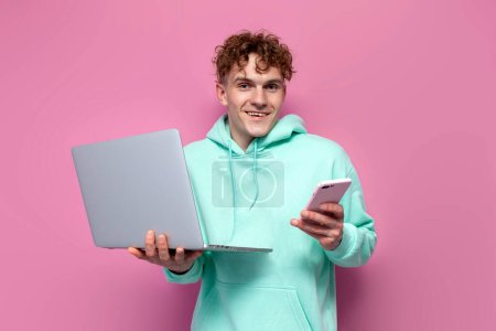 Photo for Young curly guy student in mint hoodie holds laptop and uses smartphone on pink isolated background, millenial man in blue sweatshirt holds modern devices - Royalty Free Image