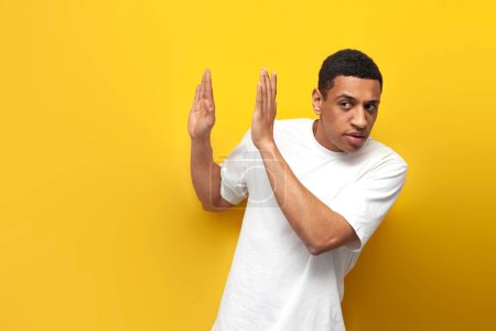 young afro american guy in white t-shirt avoids and refuses with his hands on yellow background, the man pulls back, the man defends himself and says no